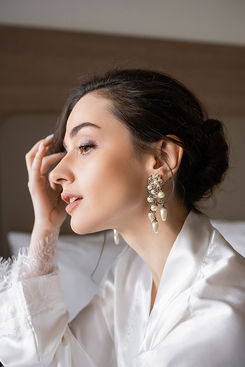 portrait of attractive woman with brunette hair in white silk robe, pearl earrings and flawless makeup preparing for her wedding in hotel room, special occasion, young bride