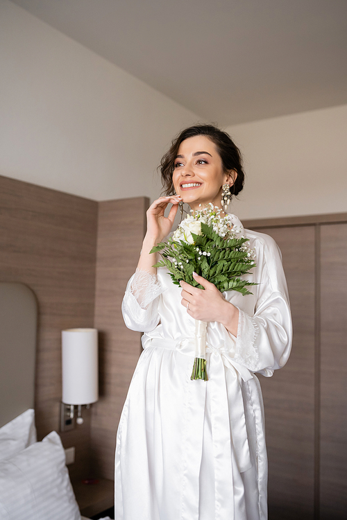 young woman with brunette hair in white silk robe and pearl earrings holding bridal bouquet while preparing for her wedding in hotel room, special occasion, happy bride