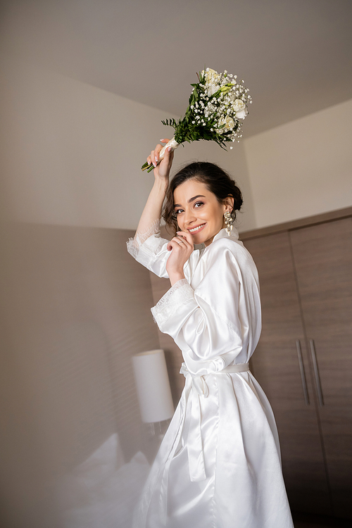 alluring young woman with brunette hair in white silk robe and pearl earrings holding bridal bouquet while preparing for her wedding in hotel room, special occasion, happy bride