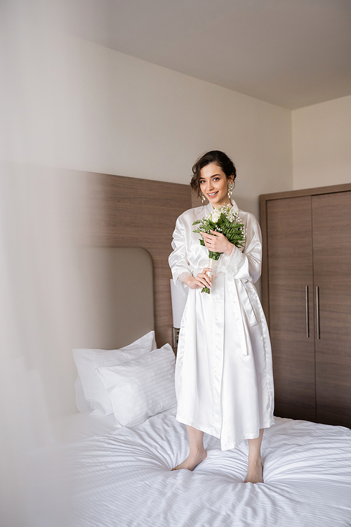 cheerful young bride with brunette hair in white silk robe and pearl earrings holding bridal bouquet while standing on bed in hotel room, special occasion, happy woman