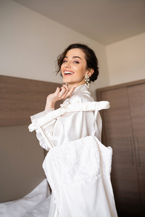 joyful bride with brunette hair in white silk robe holding soft hanger with elegant wedding dress and smiling in bedroom of hotel room, special occasion, gorgeous woman