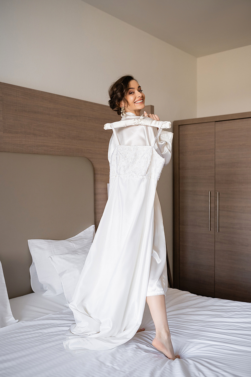 happy young bride with brunette hair in white silk robe holding soft hanger with elegant wedding dress and standing on bed in bedroom of hotel room, special occasion, pretty woman