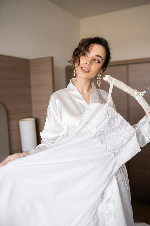 young bride with brunette hair in white silk robe holding soft hanger with elegant wedding dress in bedroom of hotel room, special occasion, charming woman