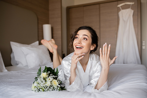 happy woman with brunette hair lying white silk robe and showing engagement ring on finger next to bridal bouquet on bed in hotel room with wedding dress on blurred background, young bride