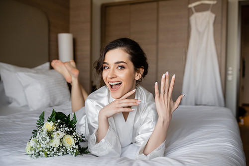 amazed woman with brunette hair lying white silk robe and showing engagement ring on finger next to bridal bouquet on bed in hotel room with wedding dress on blurred background, young bride