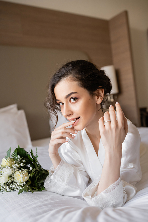 charming woman with brunette hair lying in white silk robe and showing engagement ring on finger next to bridal bouquet in hotel room on wedding day, special occasion, young bride