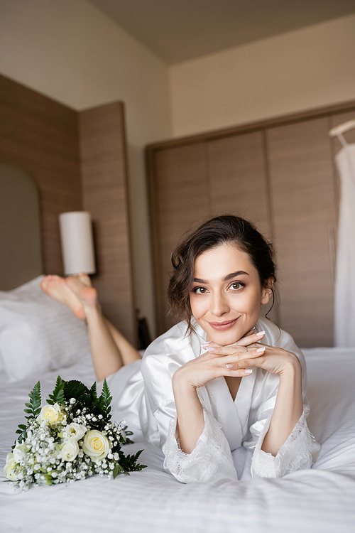 charming woman with brunette hair lying in white silk robe with clenched hands next to bridal bouquet on bed in hotel room on wedding day, special occasion, young bride