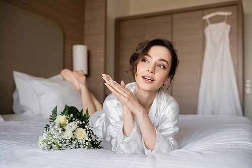 gorgeous woman with brunette hair and engagement ring on finger looking away and lying in white silk robe next to bridal bouquet on bed with wedding dress on blurred background, young bride