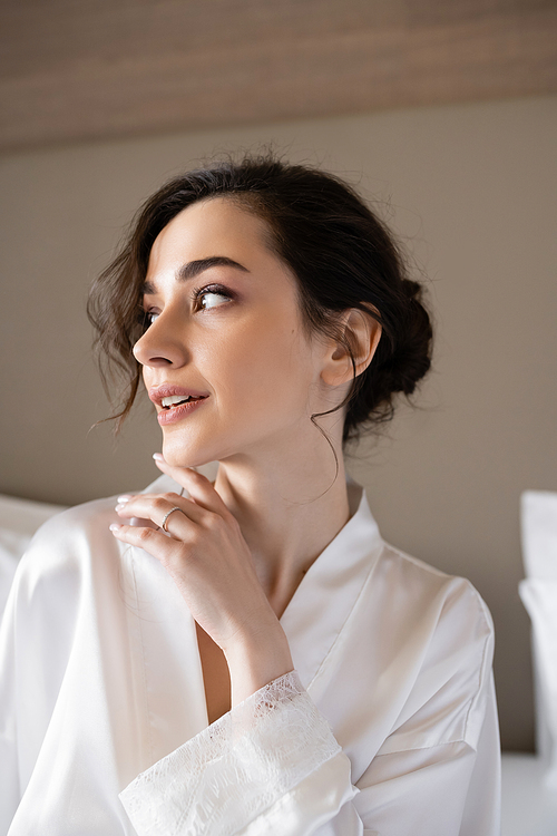 young woman with brunette hair in white silk robe showing engagement ring on finger while looking away in hotel room on wedding day, special occasion, young bride