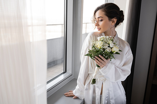 young woman with engagement ring on finger standing in white silk robe and holding bridal bouquet next to tulle curtain and window in hotel suite, special occasion, bride on wedding day