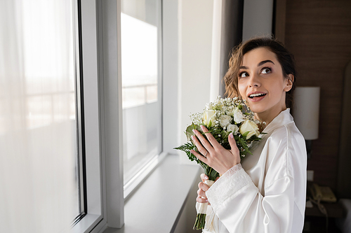 amazed young woman with engagement ring on finger standing in white silk robe and holding bridal bouquet while looking up next to window in hotel suite, special occasion, bride on wedding day