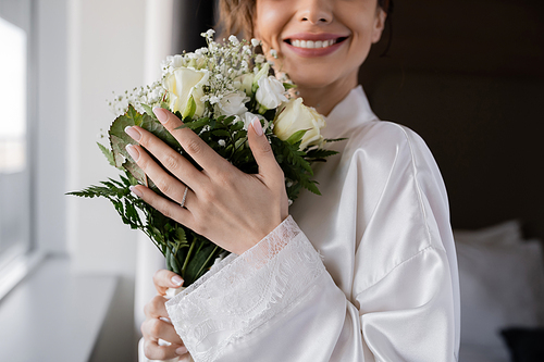 cropped view of happy bride with engagement ring on finger standing in white silk robe and holding bridal bouquet next to window in hotel suite, special occasion