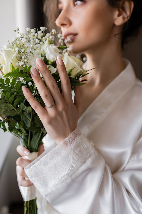 blurred bride with engagement ring on finger standing in white silk robe and holding bridal bouquet in modern hotel suite on wedding day, special occasion