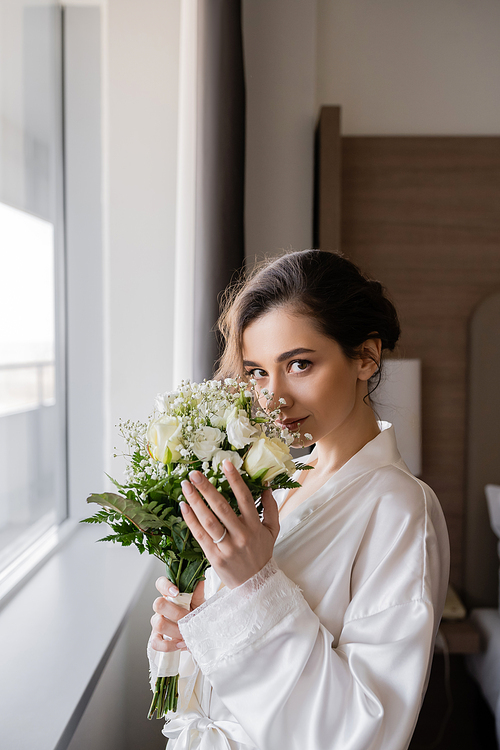 young bride with engagement ring on finger standing in white silk robe and smelling bridal bouquet next to window in hotel suite, special occasion, wedding day