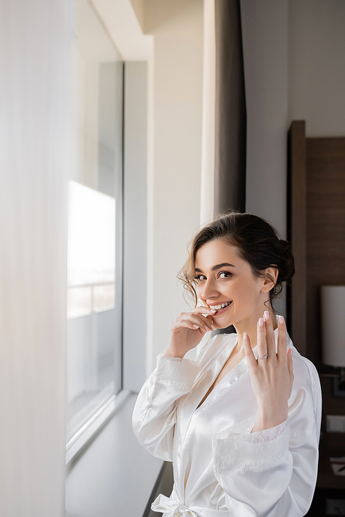 excited young bride with brunette hair in white silk robe showing engagement ring on finger and smiling next to window in hotel suit on wedding day, special occasion, hand near lips