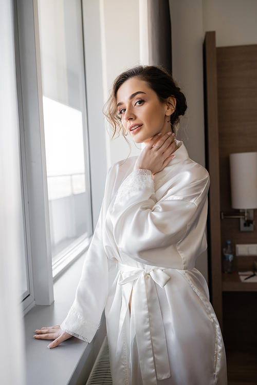 young woman with engagement ring on finger standing in white silk robe and looking at camera next to window in hotel suite, special occasion, bride on wedding day