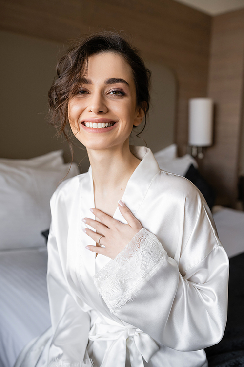 happy young bride with brunette hair and engagement ring on finger smiling while sitting in white silk robe and looking at camera in hotel room on wedding day, special occasion