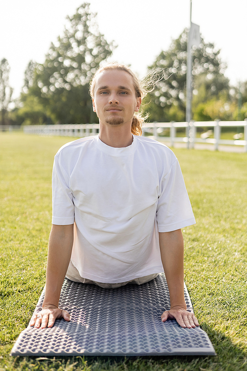 young positive man in white t-shirt looking at camera while practicing cobra pose on yoga mat outdoors