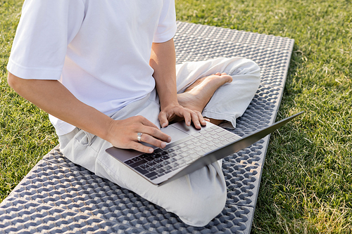 cropped view of man using laptop while sitting in easy pose on yoga mat on green grass