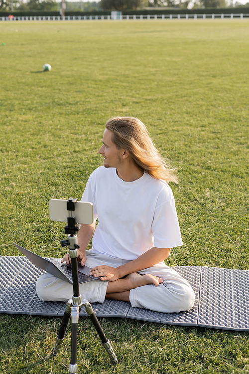 yoga coach sitting with laptop near cellphone on tripod and looking away on grassy stadium