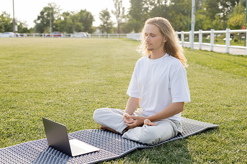 young man meditating in easy pose near laptop on yoga mat