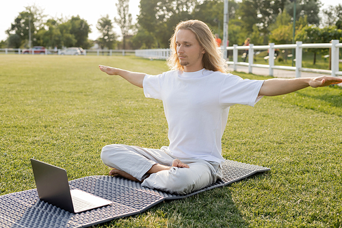young man sitting in easy pose with outstretched hands during online yoga lesson on laptop outdoors