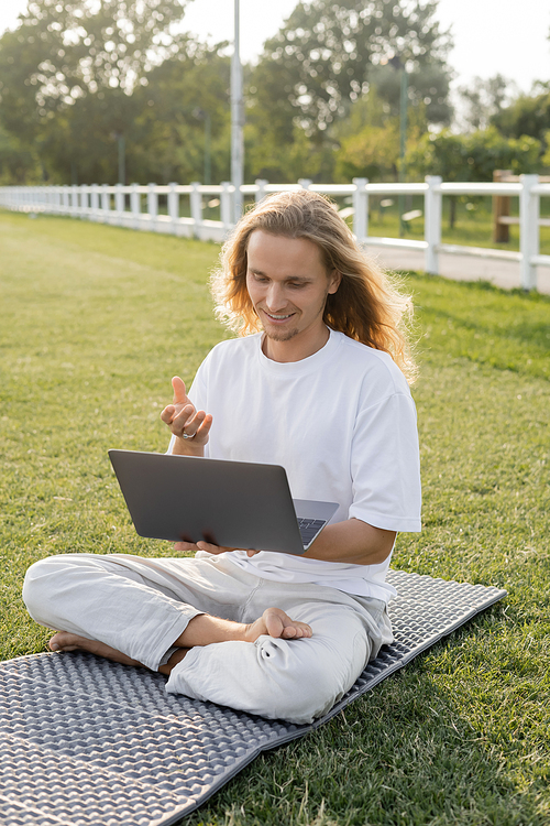 smiling man talking during yoga lesson on laptop while sitting in easy pose on outdoor stadium