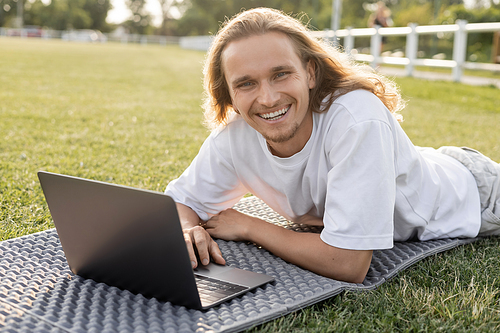 young and carefree man looking at camera while lying near laptop on yoga mat