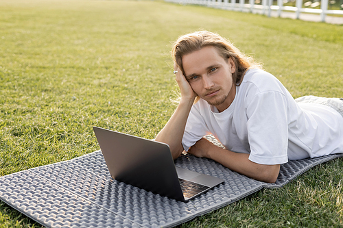 dreamy yoga man looking at camera while lying near laptop on green grass outdoors