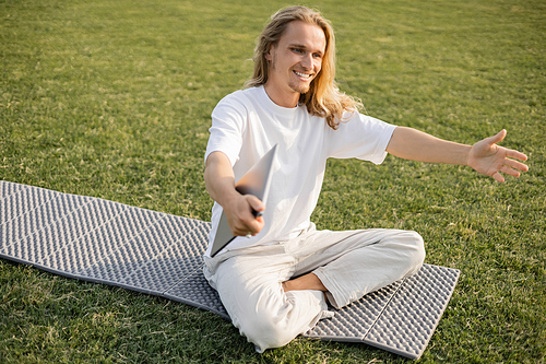 overjoyed man with laptop showing greeting gesture while sitting on yoga mat on green lawn outdoors