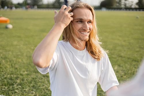 young and carefree yoga man touching long hair and looking away outdoors on blurred foreground