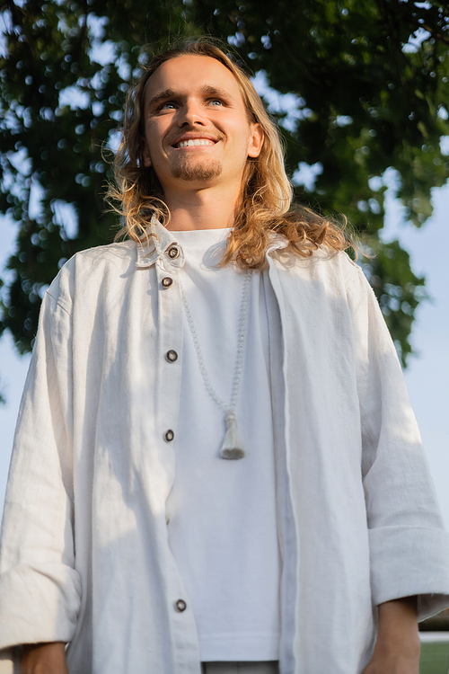 low angle view of carefree long haired yoga man in linen clothes smiling outdoors