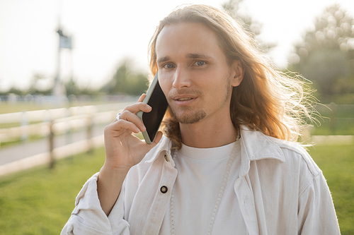 stylish long haired yoga man looking at camera during conversation on mobile phone outdoors