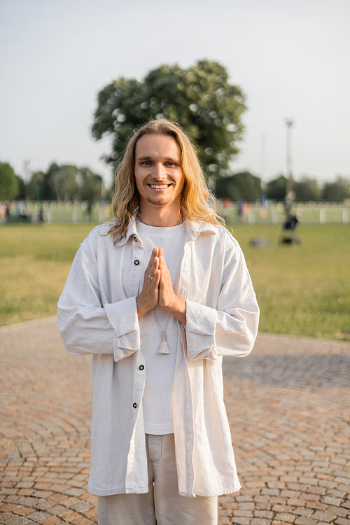 long haired yoga man looking at camera and showing anjali mudra gesture while standing outdoors