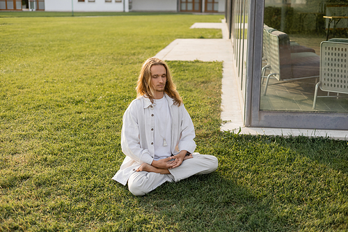 stylish long haired man meditating in lotus pose while sitting on grassy lawn near building