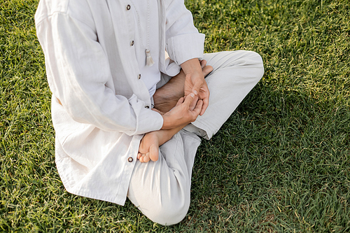 partial view of barefoot man in white linen clothes meditating in lotus pose on green grassy lawn