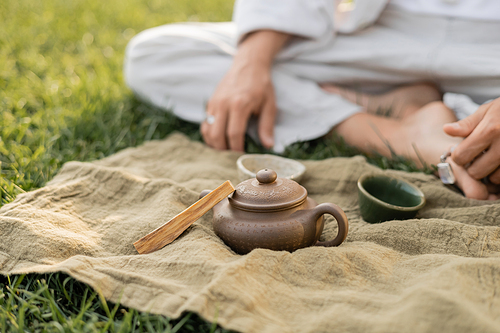 partial view of yoga man sitting near linen rug with palo santo stick and clay teapot with cups on green lawn