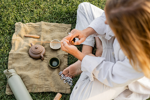 top view of yoga man holding scented stick near linen rug with clay teapot and bowls on grassy lawn