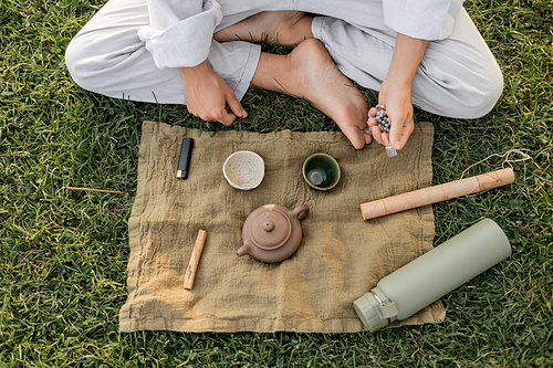top view of cropped barefoot man in white clothes holding mala beads near linen rug with ceramic cups and teapot on green lawn