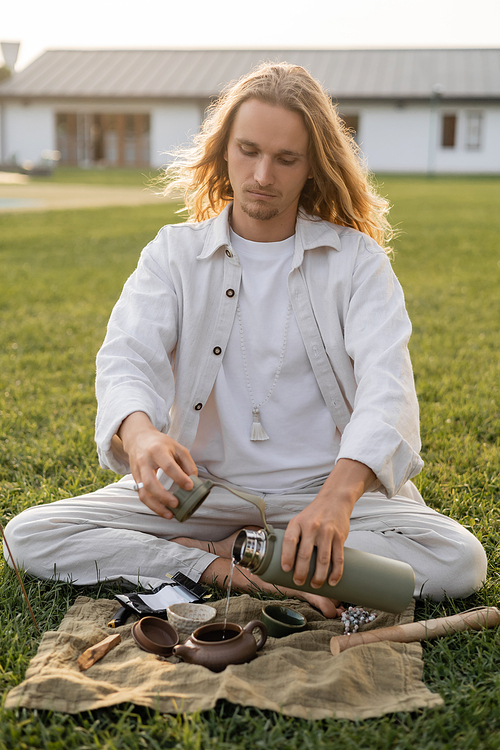 long haired man in linen clothes pouring hot water from thermos into ceramic teapot