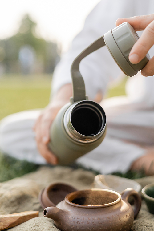 partial view of blurred man holding thermos near clay teapot during tea ceremony outdoors