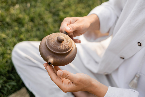 cropped view of man in white linen clothes holding clay teapot while sitting outdoors