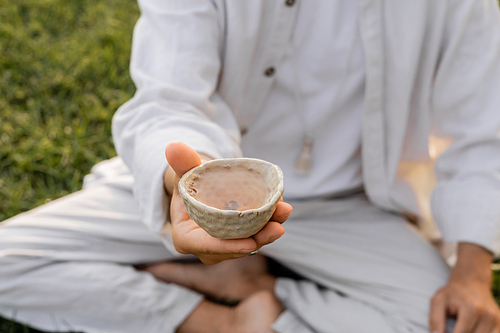 partial view of yoga man in white clothes holding clay cup with tea outdoors