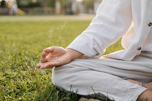 partial view of man in white clothes sitting in easy yoga pose and meditating near fragrant smoke on grassy lawn