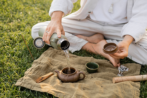 partial view of yoga man pouring hot water in clay teapot while sitting on lawn near linen rug with mala beads and palo santo stick