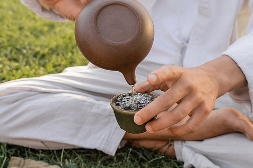 partial view of yoga man in linen clothes sitting on green lawn and pouring puer tea from oriental teapot in clay bowl