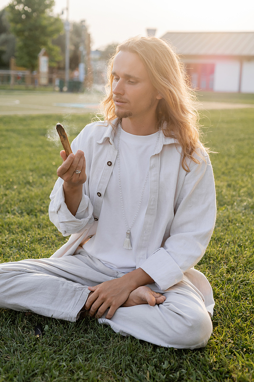 stylish man in white linen clothes sitting in easy pose and holding smoldering palo santo stick while meditating outdoors