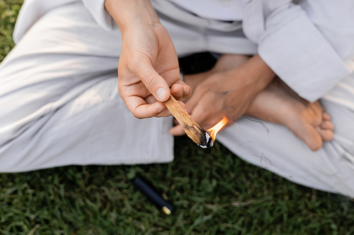 top view of cropped man holding burning palo santo stick while sitting in easy yoga pose outdoors