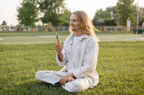 happy long haired man holding aromatic palo santo stick while meditating in easy pose on green lawn