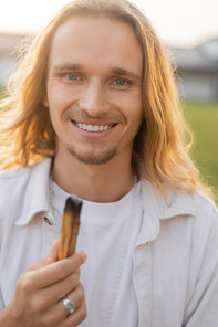 joyful long haired yoga man holding smoldering palo santo stick and smiling at camera outdoors